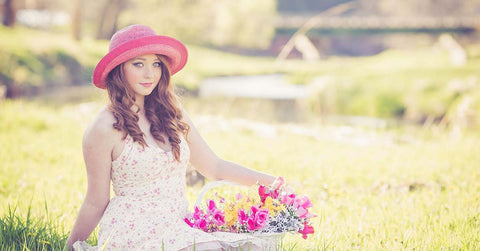 Woman sitting in a grassy field wearing pink hat, white dress holding colourful flowers