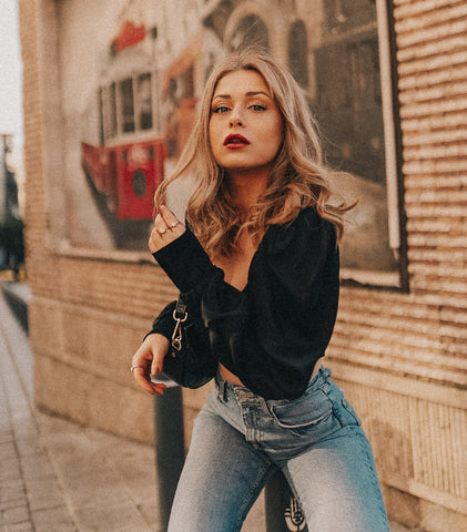 Tanara pozand in jeans si crop-top asezata pe un stalpisor metalic, in fata unui zid cu banner urias cu tramvai