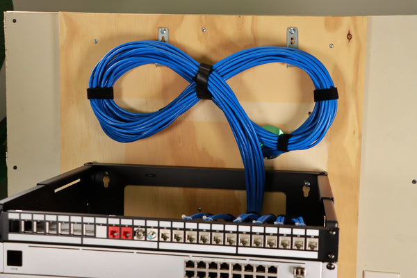 Image of a pair of blue cables hanging on a wooden wall. The cables are connected to a patch panel. The text "trueCABLE" is visible on the cables.