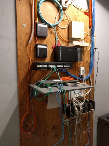 Image of a wall with a bunch of wires on it. The wires are connected to a patch panel and a router.