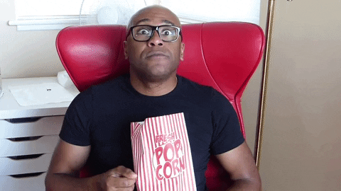 popcorn guy gif, man with glasses sitting on a red chair, entertained and shoving popcorn in his mouth
