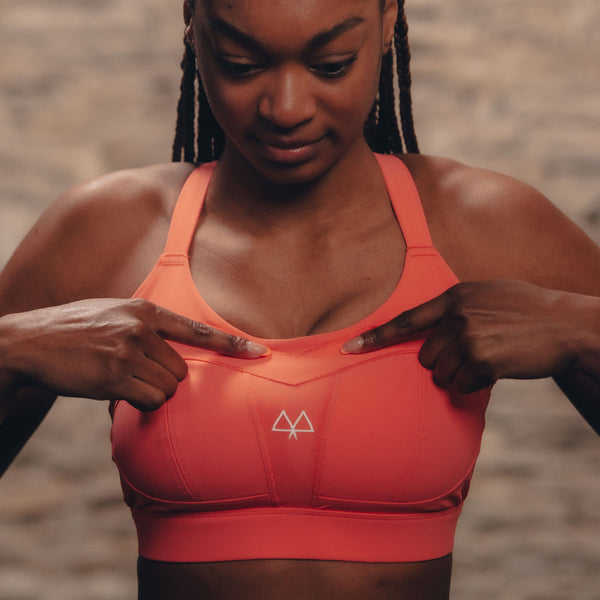 Torse shot of a young girl pointing with both hands to a coral sports bra she is wearing whilst standing in front of a light brick background