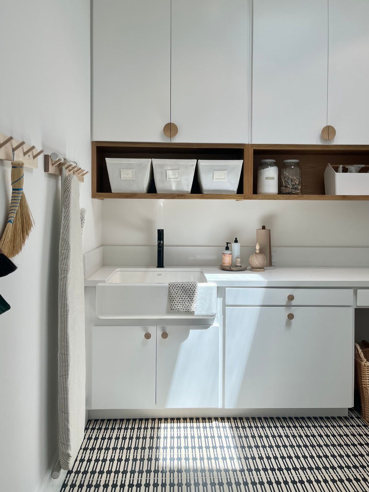 A laundry room with modern white cabinets, apron front sink and a patterned black and white tile floor.