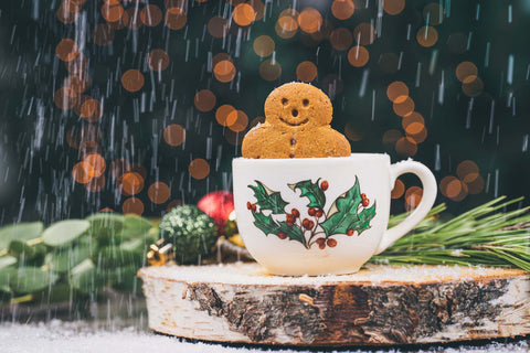 Gingerbread man in a white mug with a holly leaf and berry printed on the mug. on the piece of wood with pine in the background and out of focus lights and snow falling in the background.