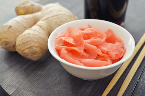 A bowl of pink pickled ginger with chop sticks and a fresh ginger root sitting next to it.