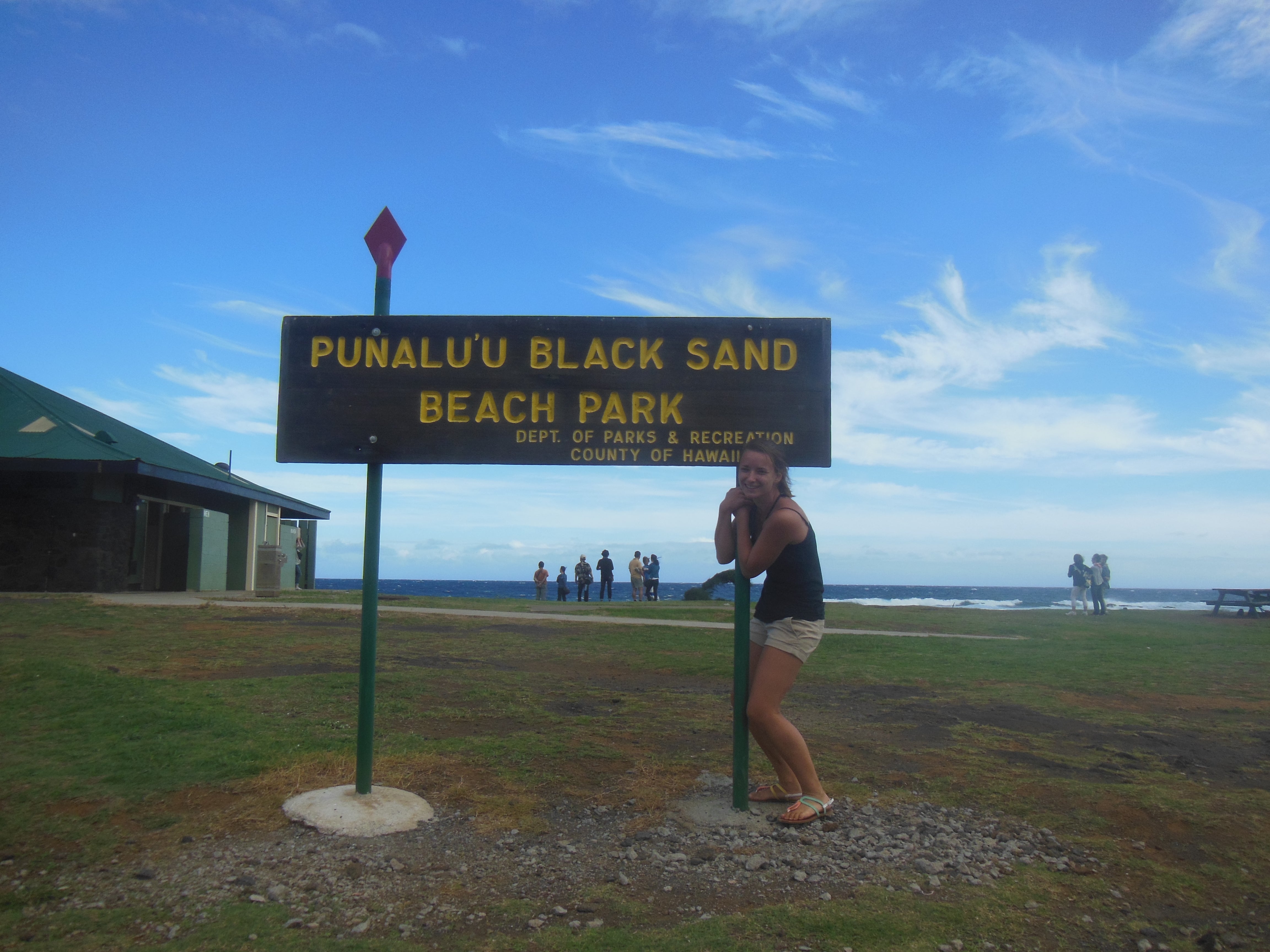 The image shows a woman holding on to the sign of Punaluu Black Sand Beach Park on the Big Island of Hawaii