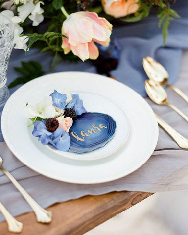Agate place card holder with gold script. On top of white plate and light blue linen tablecloth. Gold cutlery and flowers.