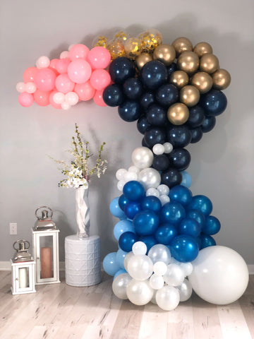 White, gold confetti, gold, light blue, dark blue and pink balloon garland against a gray wall.