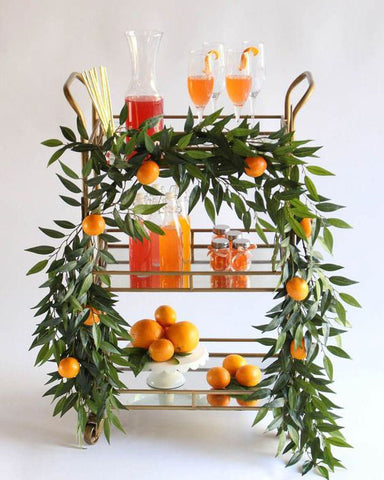 Bronze bar cart with fruit juices. Gold straws, glassware, carafe, fresh fruit, white cake stand and garland around the cart.