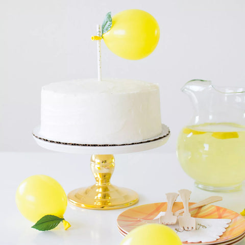 Vanilla cake on top of cake stand plate with a gold base. Small yellow balloon placed as a cake topper to mimic a lemon.