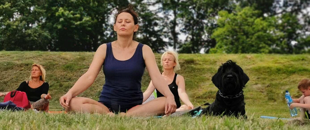 3 white women and one black dog sit on a green hillside in a yoga class