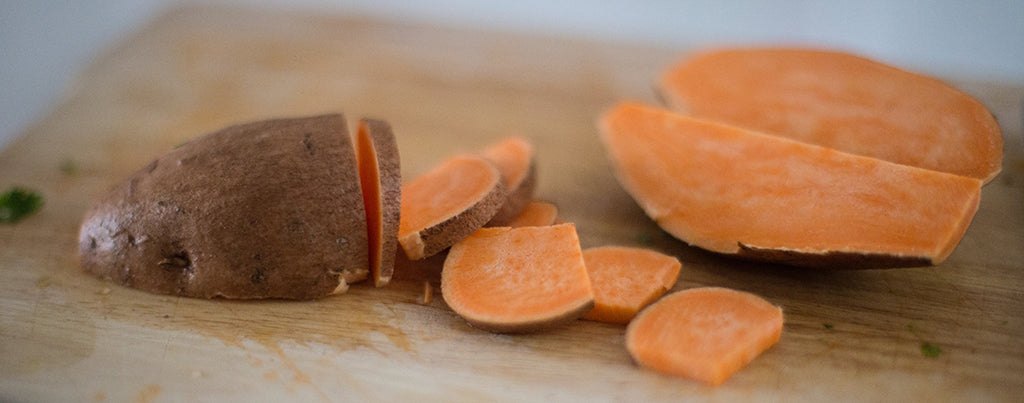 half a sweet potato sits on a wooden chopping board, the other half is sliced.