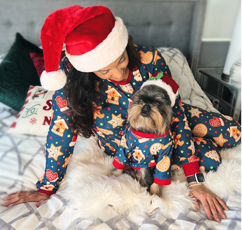 A dog and their owner sit side-by-side in bed together, wearing matching pajamas and Santa Hats!
