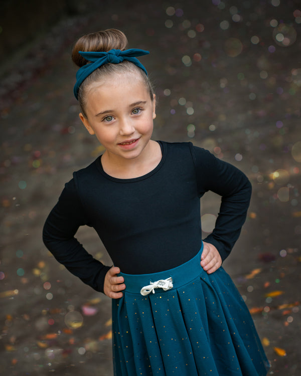 Girl wearing a teal headband and black long-sleeve tee tucked into a teal tulle skirt. 