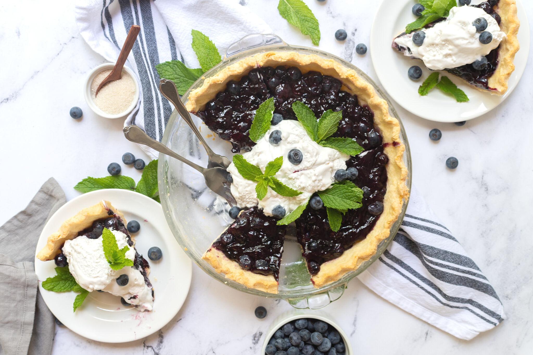 Beautiful flat lay bird-eye shot of a delicious fresh blueberry pie with two pieces cut out from it which are placed on separate plates next to the pie. The pie is topped with freshly whipped cream and mint leaves.