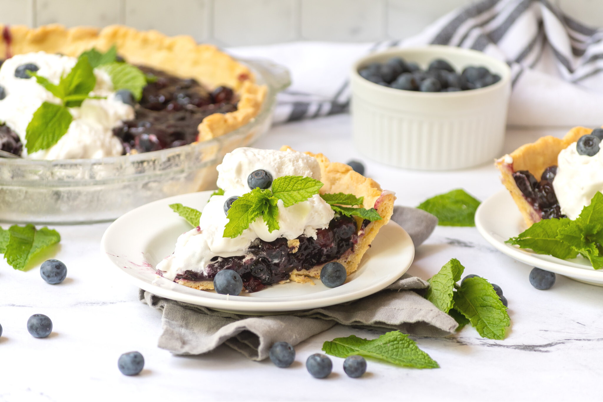 A close up shot of a piece of Blueberry pie. The pie is oozing with delicious fresh berries and is topped with freshly made whipped cream and mint leaves. In the background you can see the rest of the pie as well as a small bowl filled with fresh blueberries.