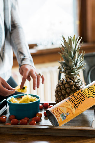 A bowl of yellow pineapple salsa, next to a pineapple, some tomatoes and a tube of Aged white cheddar chips. Someone is scooping some salsa onto a chip