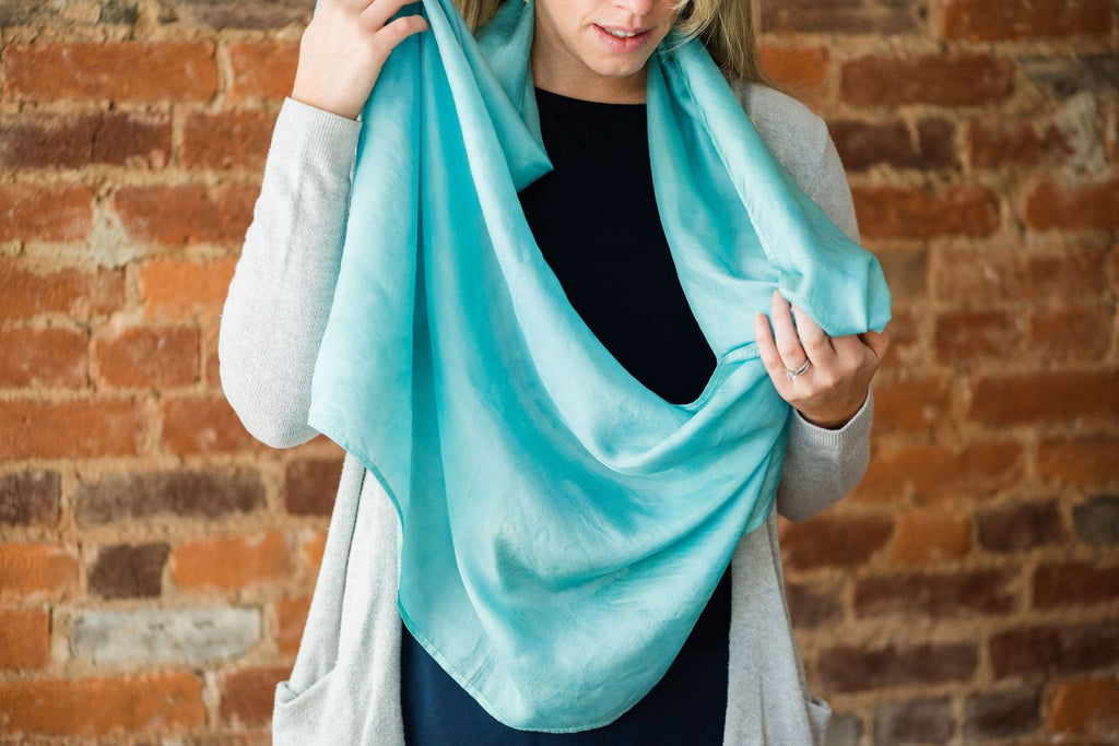 Cropped photo of a white woman wearing a light gray open cardigan and black shirt, from the nose down to the hips.  She has an aqua blue extra large silk infinity scarf wrapped once around her neck and is holding some of the fabric out in front of her in preparation to wrap the scarf around a second time.