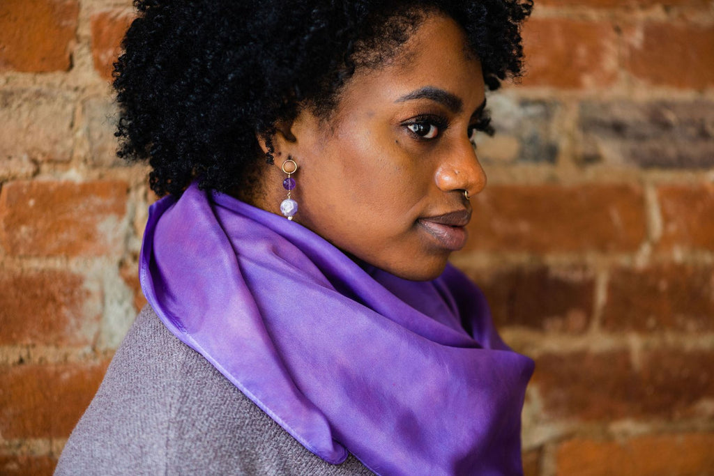 30-something Black woman in profile, wearing our amethyst earrings and crocus silk scarf.  Her dark hair is in coiling curls around her head and she's looking at the camera from the side.
