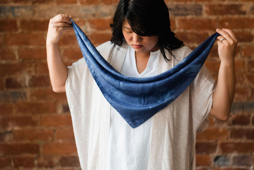 Chinese American woman with long black hair falling behind her shoulders.  She's wearing an oatmeal-colored short sleeve sweater over a white blouse and holding a midnight blue silk bandana in front of her by opposite points, so it falls into a triangle.