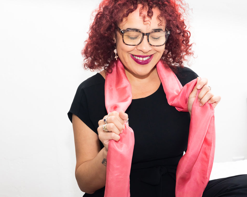 Light skinned black latinx woman with curly hair dyed red, wearing dark frame glasses, a black t-shirt, pearl earrings & rose silk scarf. She's smiling and looking down while holding the long ends of the scarf up with each hand.