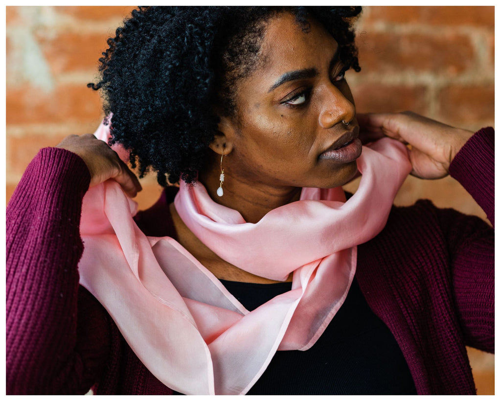 Black woman with short, full hair in curls wearing a magenta open front sweater, black shirt and jeans putting on an11x76 inch silk infinity scarf in Shell Pink