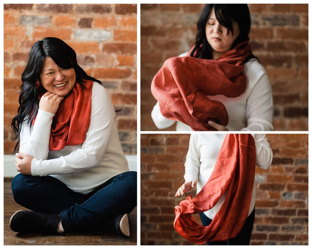 Photo collage of Chinese American woman with long, wavy hair, wearing a white pullover sweater and Russet red extra large silk infinity scarf.  In the first photo she's sitting cross-legged on the floor and grinning.  In the second, her face is blurred out in the back as she raises the scarf up to put it on.  In the third photo you see her lower torso and legs, and her hand dropping one end of the scarf.