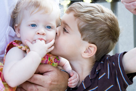 Photo of a baby girl sucking on two of her fingers. Her older brother is giving her a kiss on the cheek.