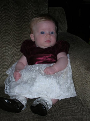 Photo of Katie at 3 months old, taken in November. She is wearing her first Christmas outfit which is a dress with a dark red velvet bodice and red satin bow with a lace skirt. She is also wearing lace ruffled socks and black patent Mary Jane shoes, sitting up on a brown couch.