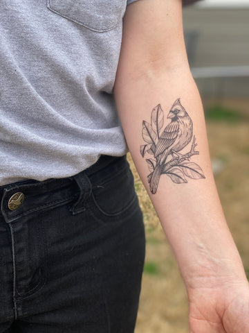 persons arm held out showing a black lined and stippled temporary tattoo of a cardinal bird sitting on a branch