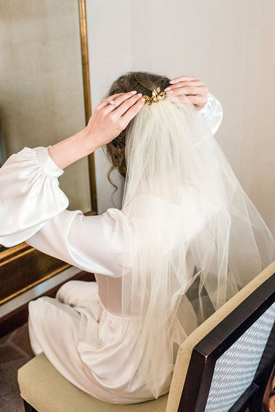 Bride wearing a white silk satin robe, putting on her wedding veil