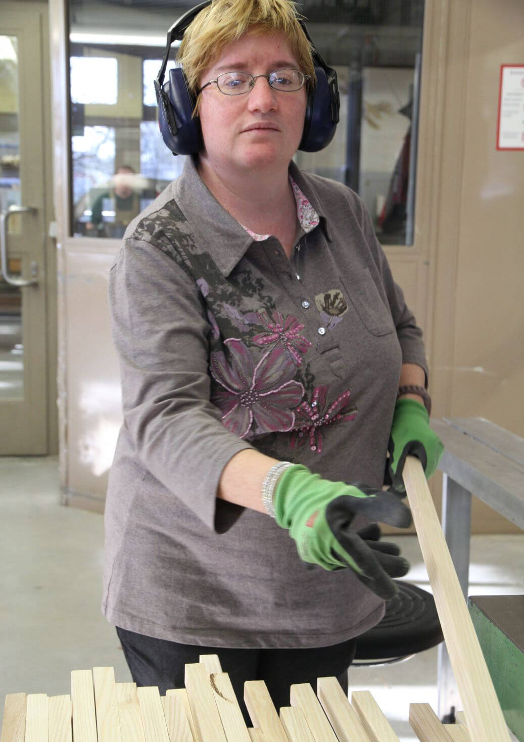 A female Metalog Tools employee wearing green gloves selects a raw birch wood board to be handcrafted into a team building tool