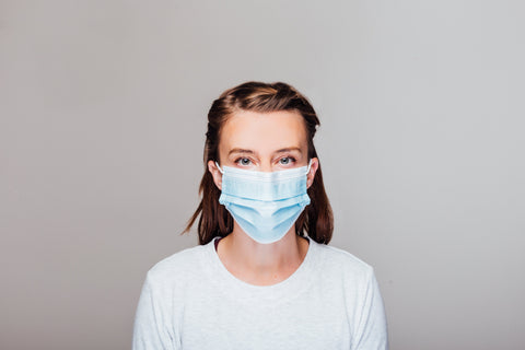 Woman in a white t-shirt stands in front of a grey background, wearing a blue disposable mask