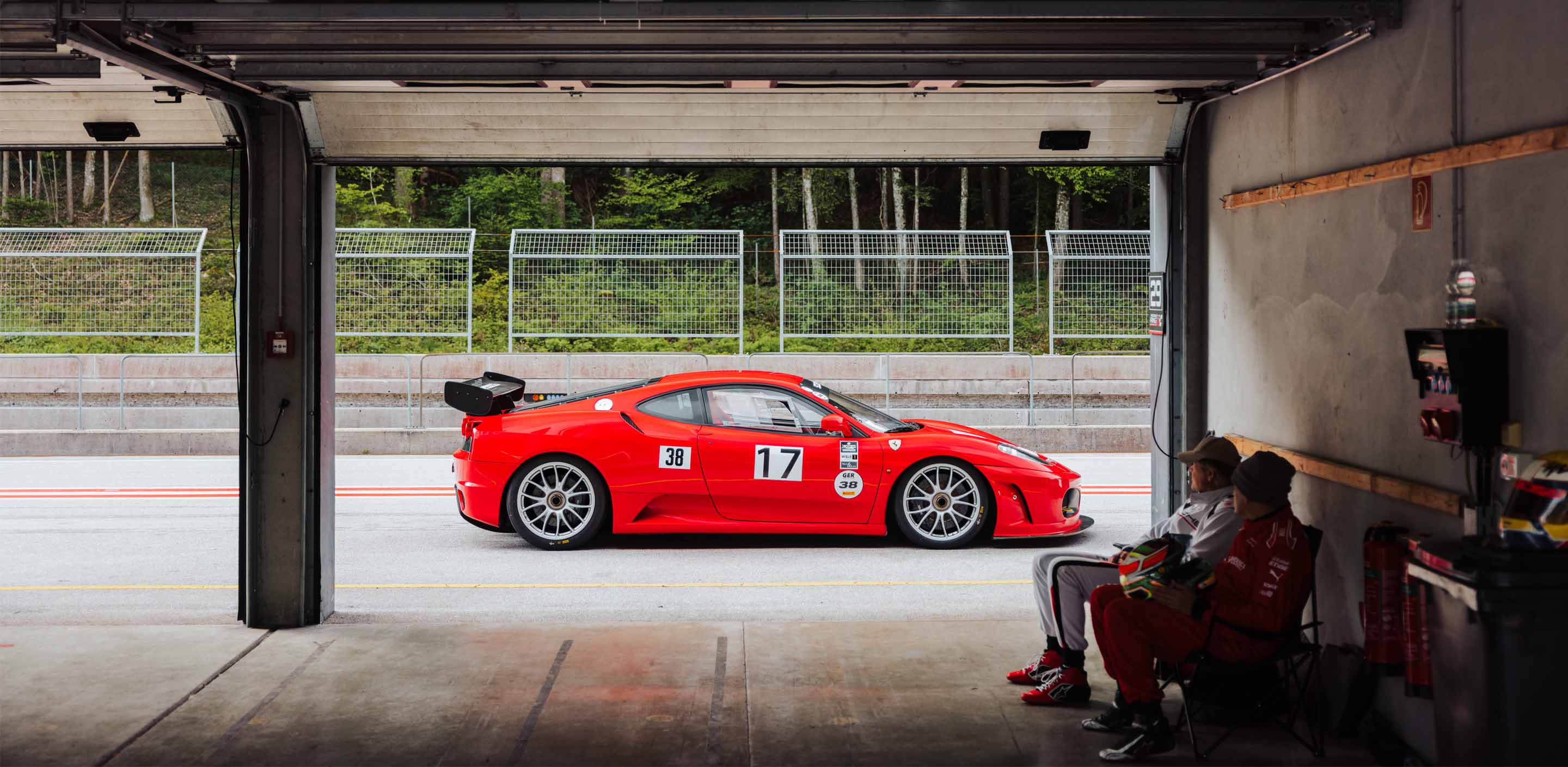 View on a Ferrari 360 Challenge race car through a Pitbox at Salzburgring during a GP Days Open Pitlane Track Day with two driver waiting in the Box sitting on chairs.
