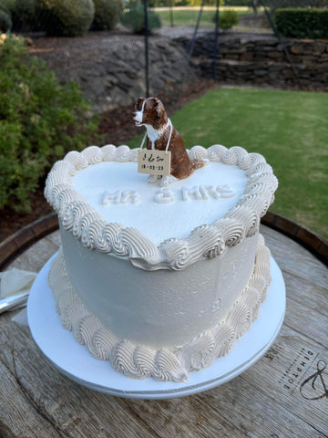 Dog cake topper on top of a wedding cake with a sign around his neck saying i do and the wedding date