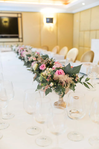 bouquets of flowers sat in a row on a long dining room table with glassware set out and a white table cloth