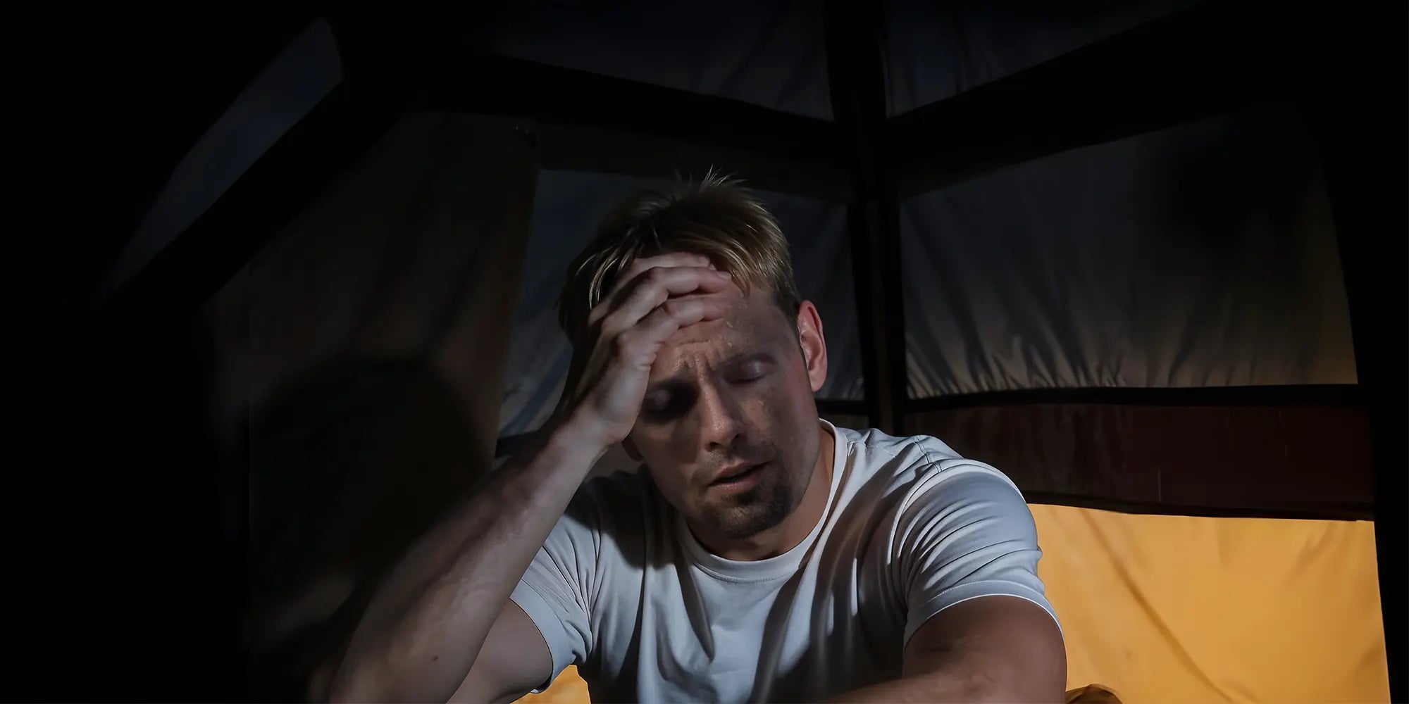 A man is sitting in a dimly lit tent at night, visibly sweating and exhausted, holding his head in discomfort, depicting the struggle of sleeping in hot, unventilated camping conditions.