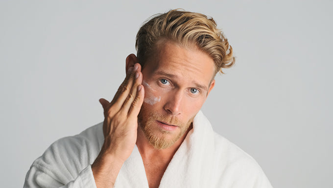 a bearded man in a robe applying moisturizer to his face