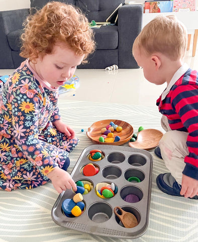 Two small children playing with loose parts and a muffin tray.