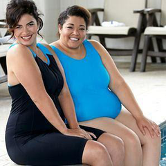 Two plus size women sitting at the edge of an indoor pool with their feet in the water. They are both smiling at the camera. They are wearing JunoActive plus size swimwear, the Aquatard and one-piece tank suit.