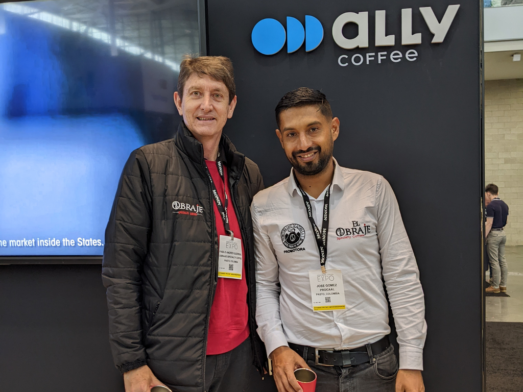 Two men standing in front of a black background with an Ally Coffee logo and part of a TV screen visible