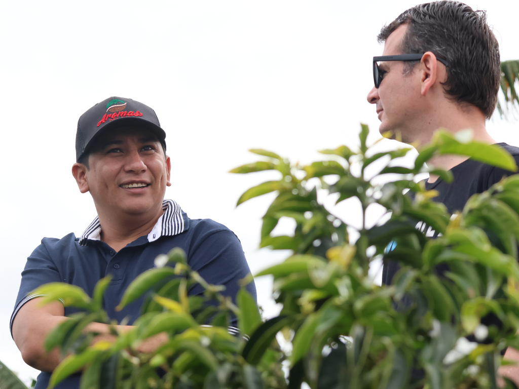 Two people talking in front of a cloudy sky with green coffee plants in the foreground