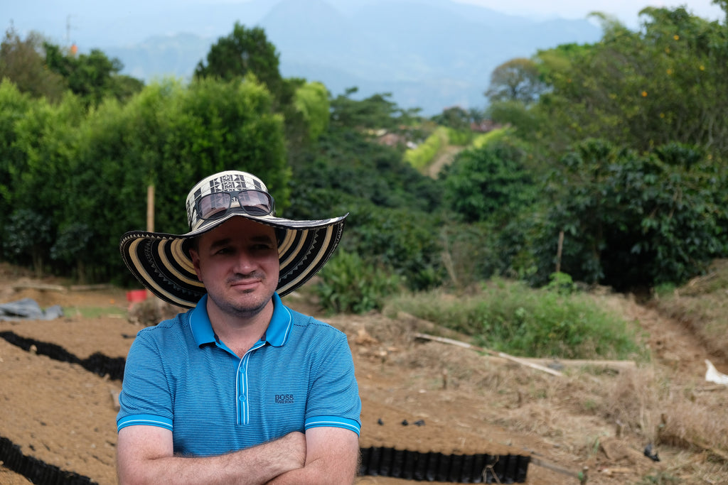 A coffee producer with his arms crossed wearing a blue polo and wide brimmed hat