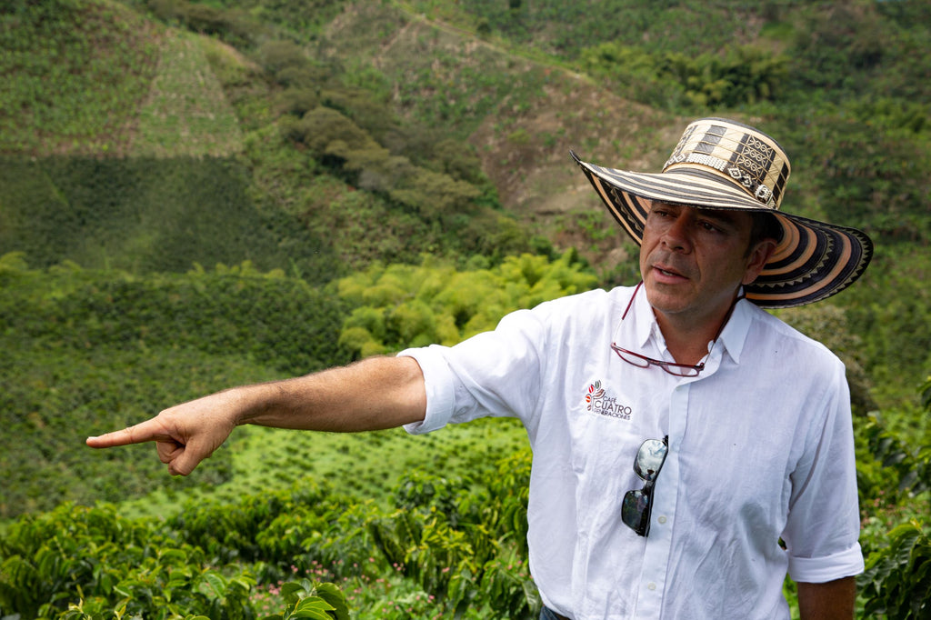 A coffee producer pointing to something out of frame, wearing a white button up shirt embroidered with the Cafe Cuatro Generaciones logo and a wide brimmed hat
