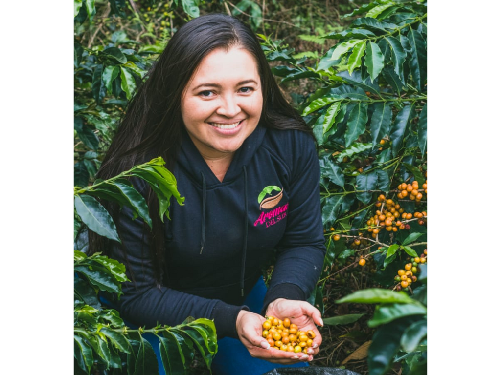 A coffee producer holding yellow coffee cherries in her hands, wearing a black hooded sweatshirt with the Aromas del Sur logo and jeans