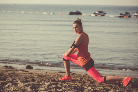 woman on beach close to waters edge wearing pink and coral performing a hip flexor stretch