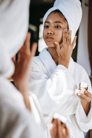 Woman adding face cream in the mirror with a towel around her head and a dressing gown on.