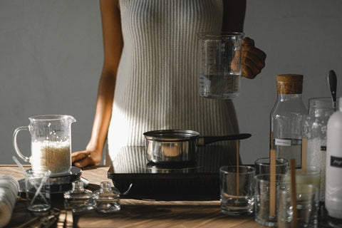 Woman standing in front of her candle making set up with a small table top stove, pot, wax in a jug and other candle making stuff