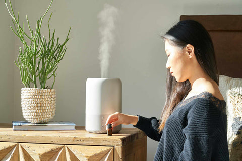 Woman touching a small amber glass bottle of essential oil that is placed on a table alongside a diffuser letting out steam and a plant
