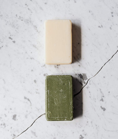 A birds eye view of two soap bars placed on a marble table one above the other. The top soap bar is white and the bottom is green. They are both rectangular.